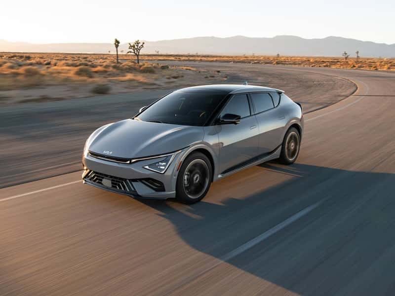 Silver 2025 Kia EV6 driving on a desert highway with trees and mountains in the distance.
