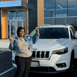 Woman in front of her new white jeep cherokee with the car dealership entrance as the backdrop