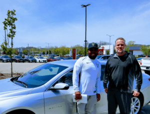 Happy Customer standing next to car salesman agasint dealership lot backdrop