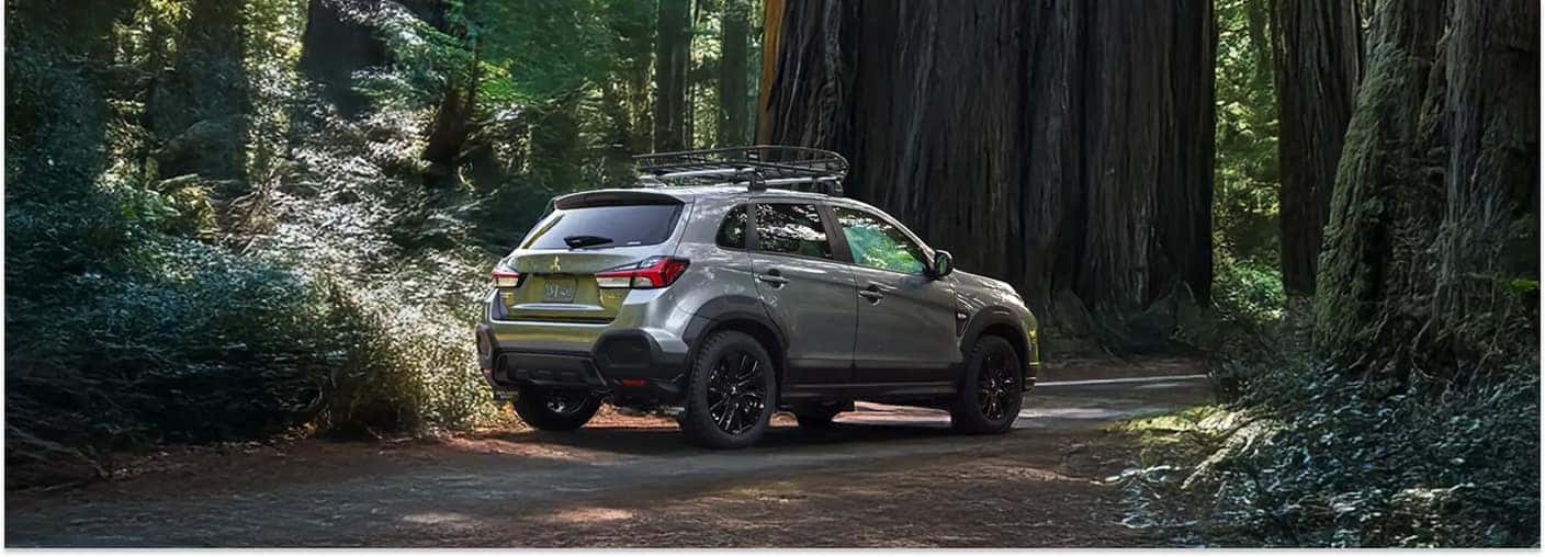 Rear three-quarter view of a silver 2026 Mitsubishi Outlander Sport with a roof rack on a forested dirt road.